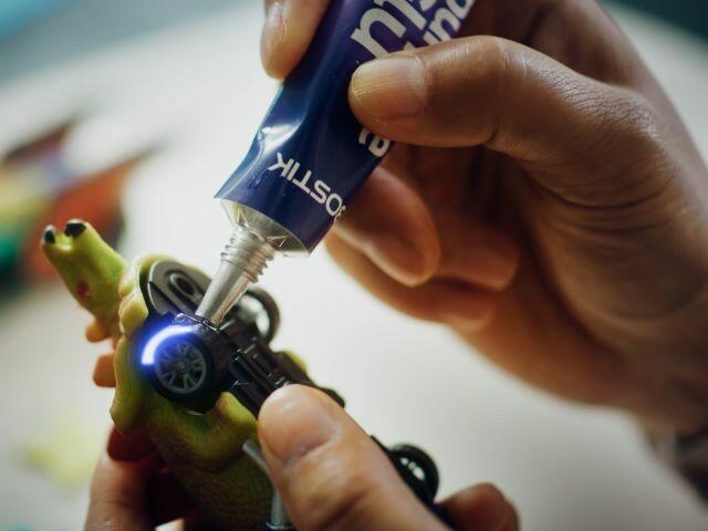 Hands using Bostik glue to repair a toy car wheel attached to a plastic dinosaur.