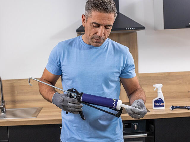 Pessoa usando pistola aplicadora para preparar tubo de selante Bostik em cozinha moderna com bancada de madeira e pia inox.