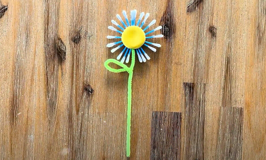 Colourful handmade spring flower made from cotton buds, a yellow bottle cap, and a green pipe cleaner stem, displayed on a wooden surface.