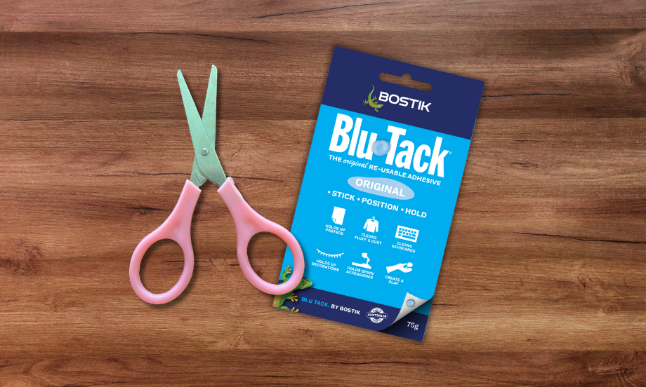 Bostik Blu-Tack pack and pink scissors on a wooden floor, illustrating the DIY floor-scratch protection tutorial.