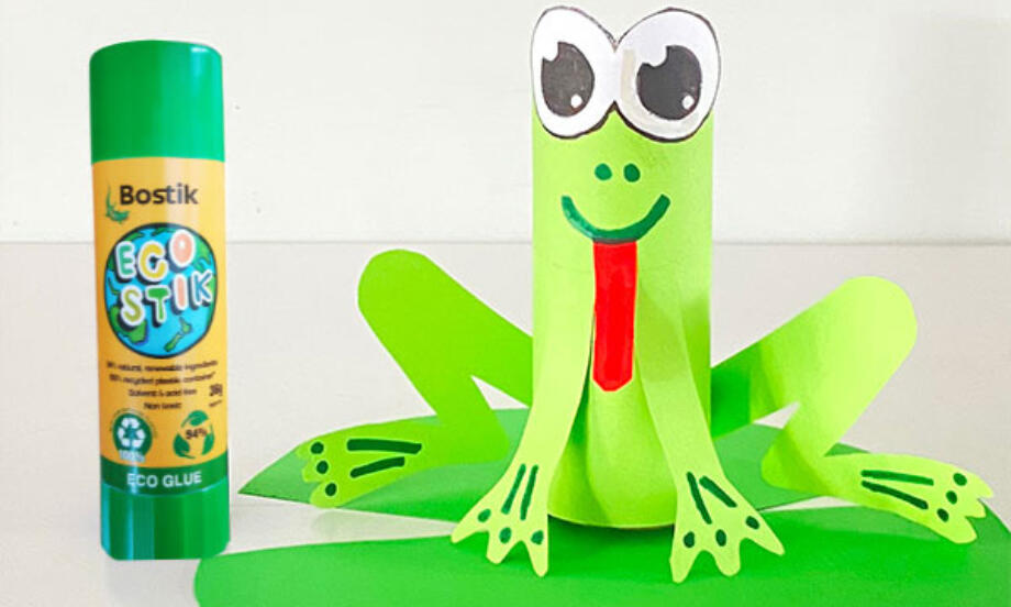 A cute paper frog craft made from a toilet roll tube, sitting on a green lily pad beside a Bostik Blu Stik glue stick.