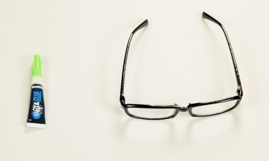 Ochelari cu rama ruptă așezați pe masă lângă tubul de adeziv Fix & Glue, pregătiți pentru reparare rapidă și întărirea îmbinării fragile.