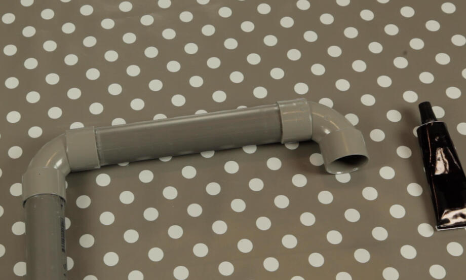 Țeavă din plastic dur cu îmbinare slăbită așezată pe masă, alături de tubul cu adeziv, sugerând pregătirea pentru o reparare rapidă și sigură.