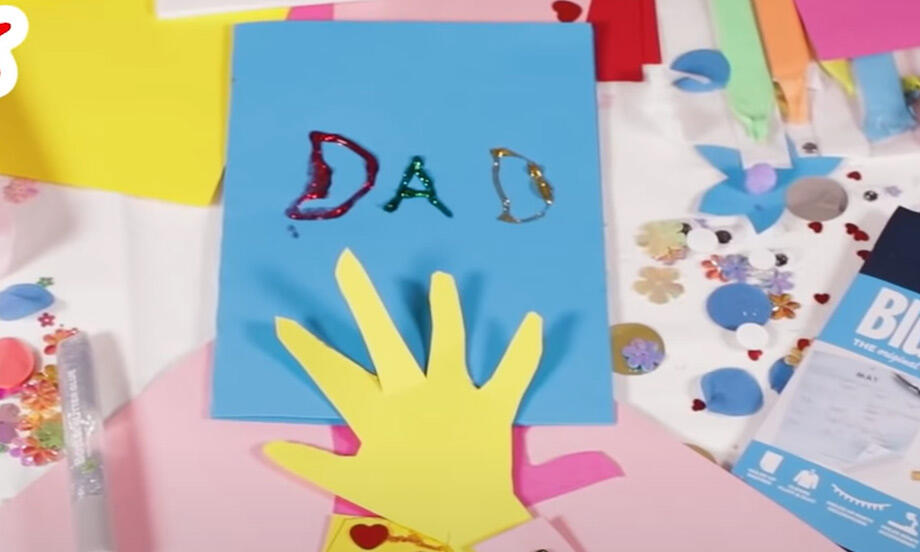A colourful handmade “Round of Applause” card holder made from yellow and pink paper hand cut-outs, holding a bright blue card decorated with glitter letters spelling “DAD”.