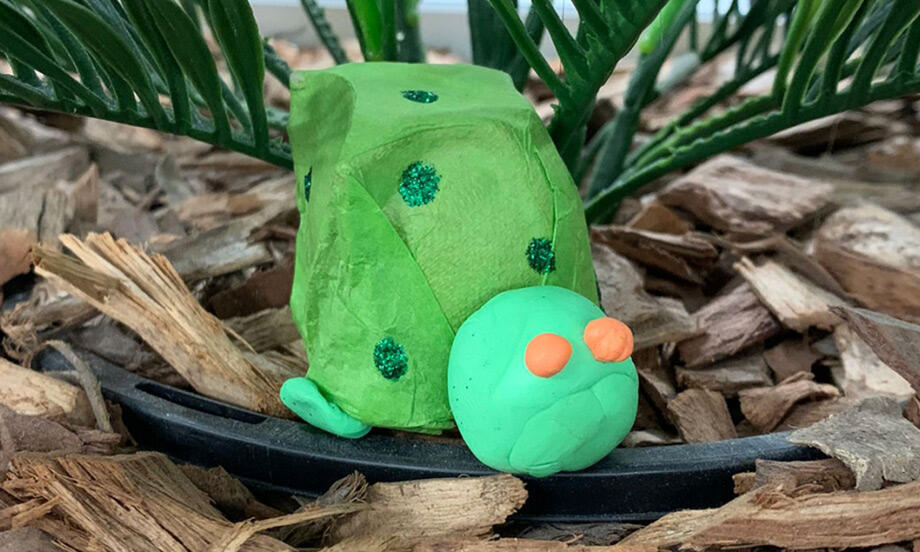 Two handmade egg carton turtles painted green with glitter spots, one with a cone shell and one with a rounded shell, placed on bark under tropical leaves.