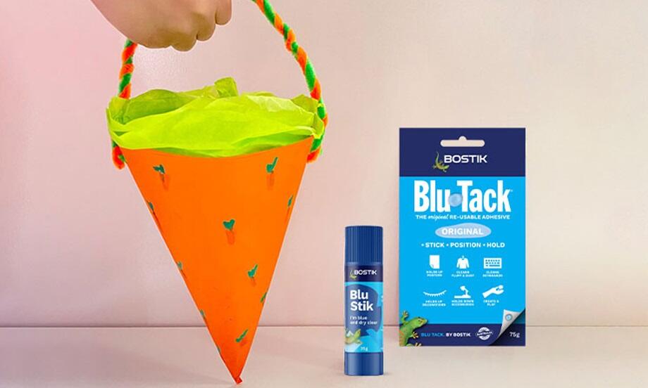 完成的橙色蘿蔔造型手作籃以綠色薄紙作內襯，搭配扭繩提把，旁放 Blu Tack 與膠棒產品，呈現復活節 DIY 籃子的趣味成品，示範簡單材料也能打造創意節日禮物容器。