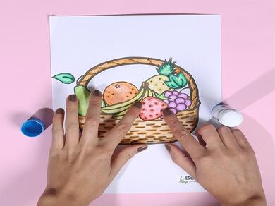 Both hands place a coloured strawberry inside the paper basket using glue, completing the fruit basket craft.