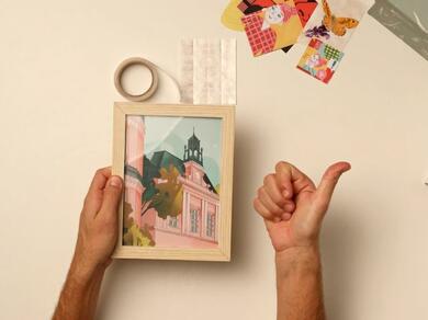 Hands holding a framed print, a roll of Glu Dots, and colourful paper pieces, ready to decorate.
