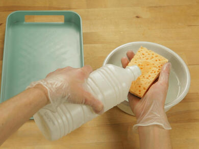 Close-up of hands pouring liquid cleaner onto a sponge above a bowl of water.