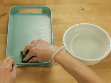 Person cleaning a green plastic tray with a sponge after glue removal.
