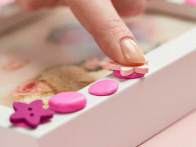 Close-up of a hand placing pink Blu Tack Colour onto a white picture frame to secure decorative buttons.