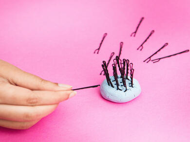 Hand inserting a hairpin into Bostik Blu Tack to organize pins on a pink surface.