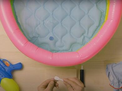 A person holding a circular adhesive patch above the hole in the side of a paddling pool.