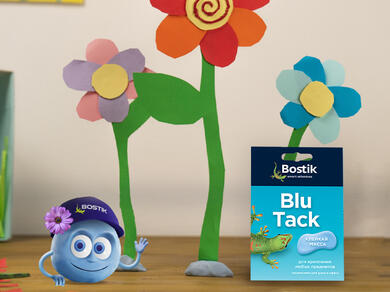 Яркие бумажные цветы стоят рядом с упаковкой Blu Tack, подчёркивая простые и увлекательные DIY‑поделки для украшения детской комнаты.