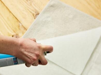 Spray glue being applied to the back of a rug for secure bonding in an alternate view.