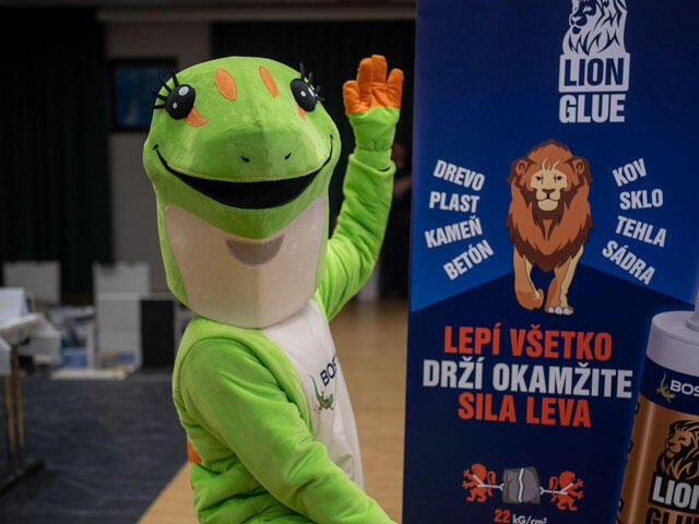 Maskot Bostik v zelenom kostýme gekona stojí vedľa bannera Lion Glue s textom o univerzálnom lepidle na drevo, kov, sklo a ďalšie materiály.