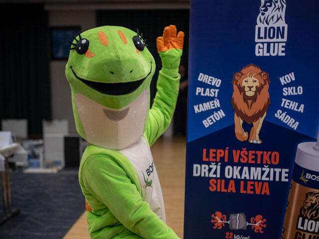 Maskot Bostik v zeleném kostýmu gekona stojí vedle reklamního banneru Lion Glue, který propaguje univerzální lepidlo pro různé materiály.
