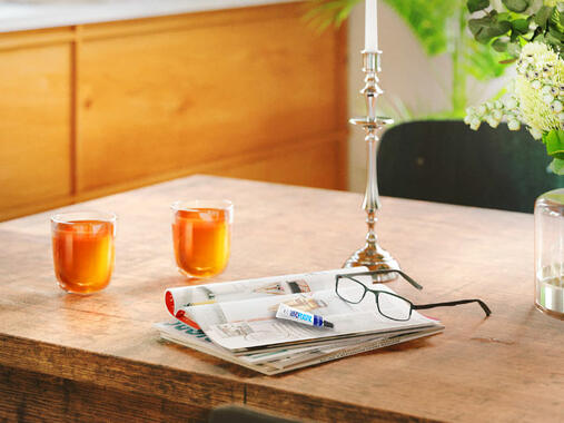 Reading glasses, hard plastics glue, and magazines on a wooden table with two glasses of iced tea