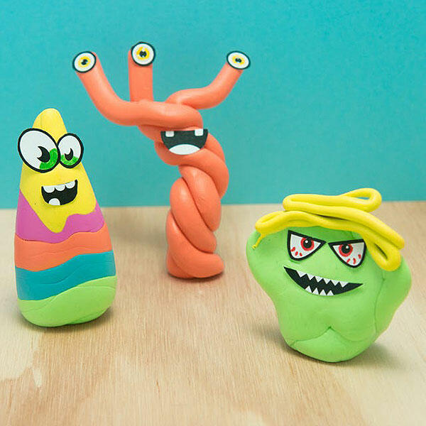 Three fun Blu Tack monsters in bright colours – one rainbow-striped with glasses, one tall and twisted with four eyes, and one green with yellow hair and sharp teeth – standing on a wooden surface against a blue background.