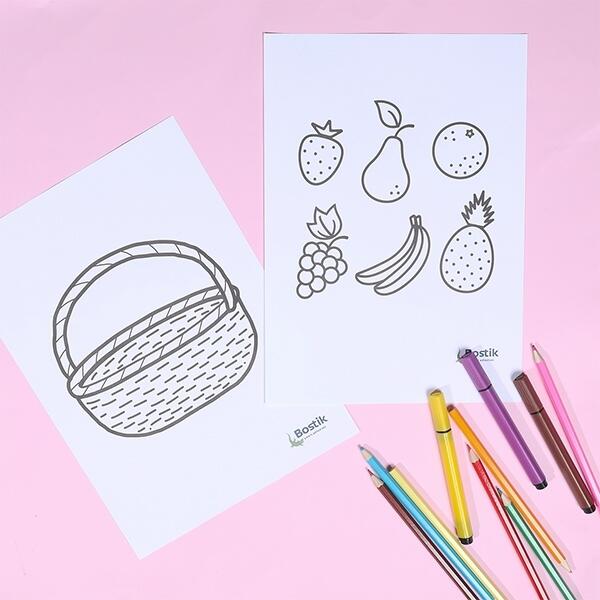 A colouring page of a fruit basket and different fruits is placed on a pink table, with coloured pencils ready to be used.