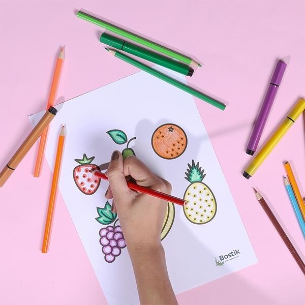 A hand colours in a strawberry on the fruit basket activity sheet using a red pencil, with other fruits already coloured.