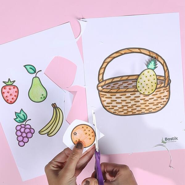 A hand cuts out an orange from the coloured fruit sheet while the pineapple is already glued inside the basket.