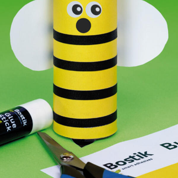 Close-up of the bee craft in progress with yellow paper wrapped around a cardboard roll, next to scissors and a Bostik glue stick.
