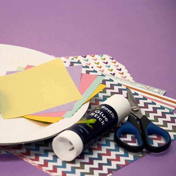 A paper plate, Bostik Glue Stick, scissors, and assorted colourful craft papers are laid out on a purple background—ready for crafting fun!