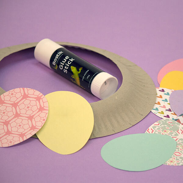 A paper plate with its centre cut out sits next to pastel-coloured egg shapes, showing the start of the wreath base.