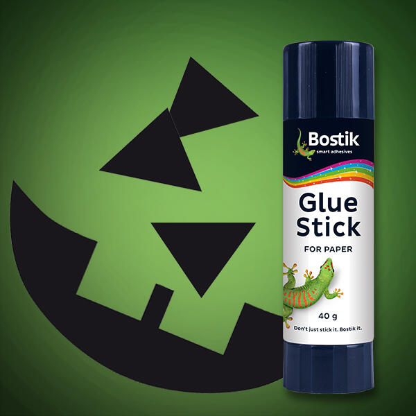 A green background with a Bostik Glue Stick and black paper cutouts of a jack-o’-lantern’s face, including triangle eyes and a smile.