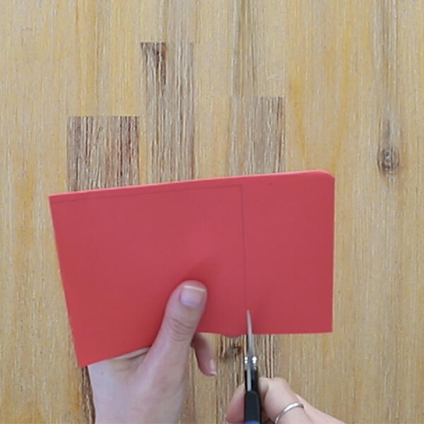 Cutting a red square piece of paper with scissors to start the paper rose craft.