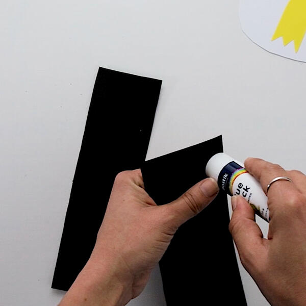 Applying glue to a black paper strip using a glue stick to start building the penguin's body.