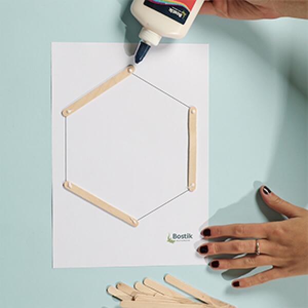 Drawing a hexagon outline on paper and placing wooden popsicle sticks along the lines to form the base shape.