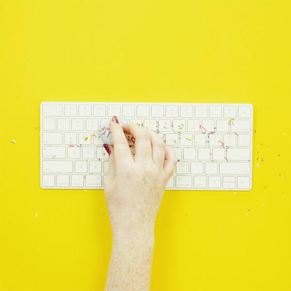 Hand using Blu Tack to lift dust and debris from a white computer keyboard on a bright yellow background.