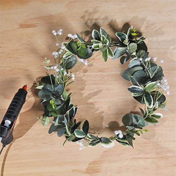 Finished DIY wreath decorated with greenery and white flowers, with a glue gun placed beside it.