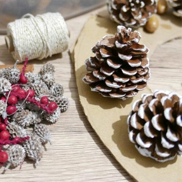 Partially assembled Christmas wreath with pinecones and acorns on cardboard next to scissors and cinnamon stick.