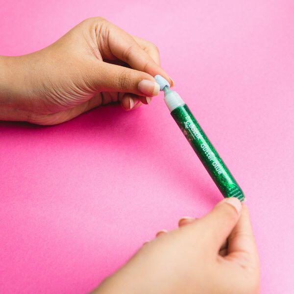 Hands applying Bostik Glitter Glue for a DIY craft project on a pink surface.