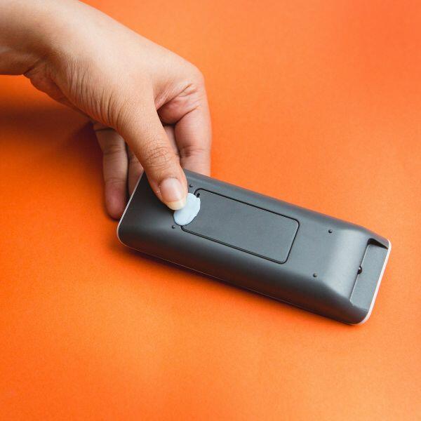 Hand attaching Bostik Blu Tack to a remote control for easy mounting on an orange surface.