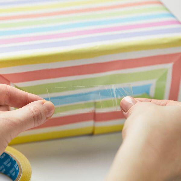 Holding a strip of Bostik Sticky Tape to wrap a colourful gift box.