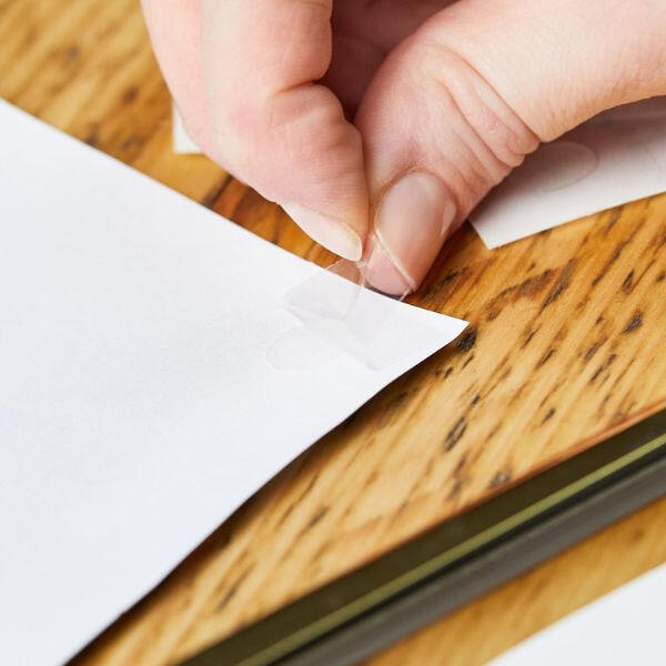 Close-up of a hand peeling an extra strong Bostik Glu Dot from white paper on a wooden table for a craft project.