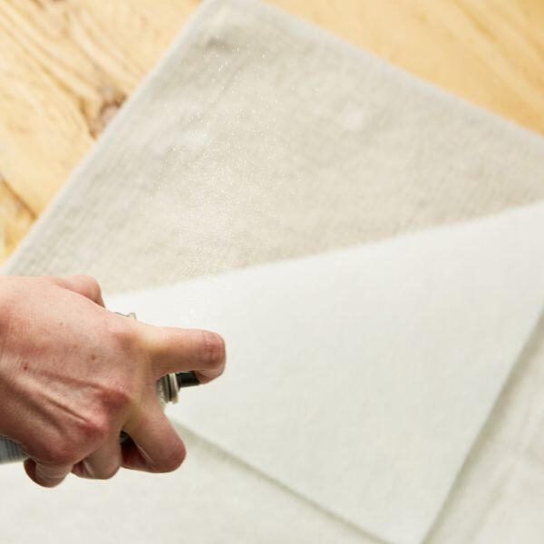 A hand spraying Bostik Spray Adhesive onto the back of a piece of white fabric over a beige surface.