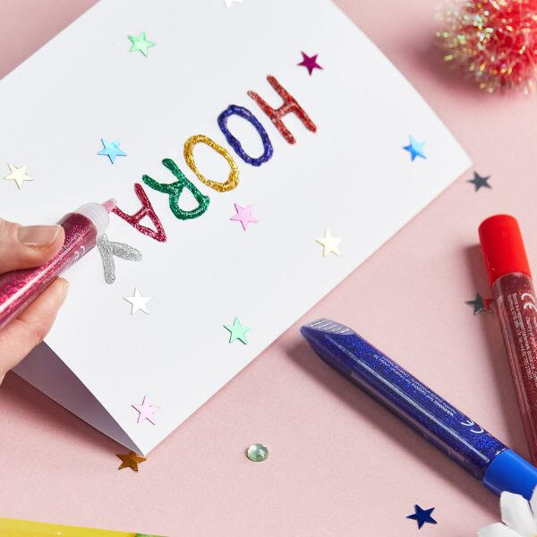 Hand using Bostik Glitter Glue to decorate a white card with the word "HOORAY" in multicoloured glitter letters and star confetti accents.