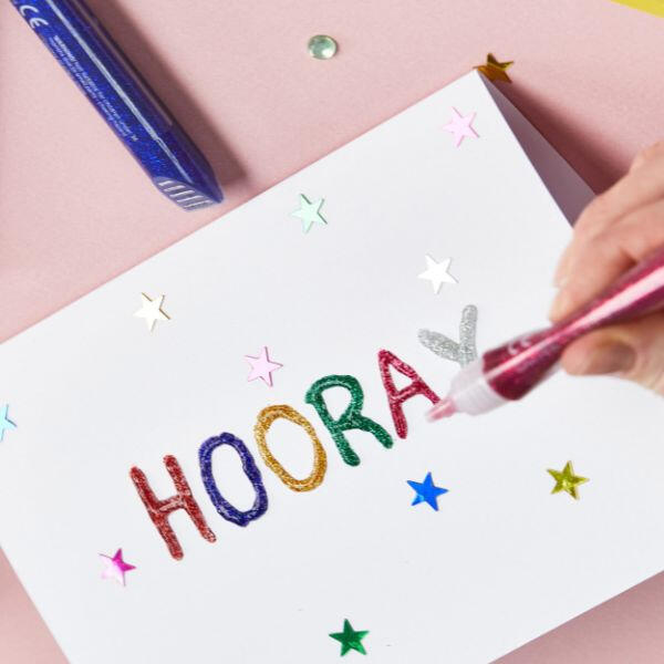 Hand using Bostik Glitter Glue to decorate a white card with the word "HOORAY" in multicoloured glitter letters and star confetti accents.