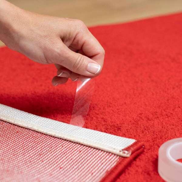Hand applying Bostik Miracle Mounting Tape to a red rug for stable floor placement.